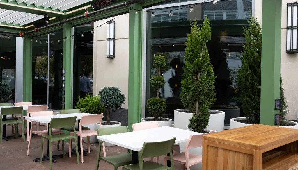 Empty restaurant patio: white tables, green/pink chairs, plants, and pergola.