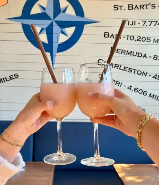 Two hands raising pink cocktails with straws, in front of a travel wall.