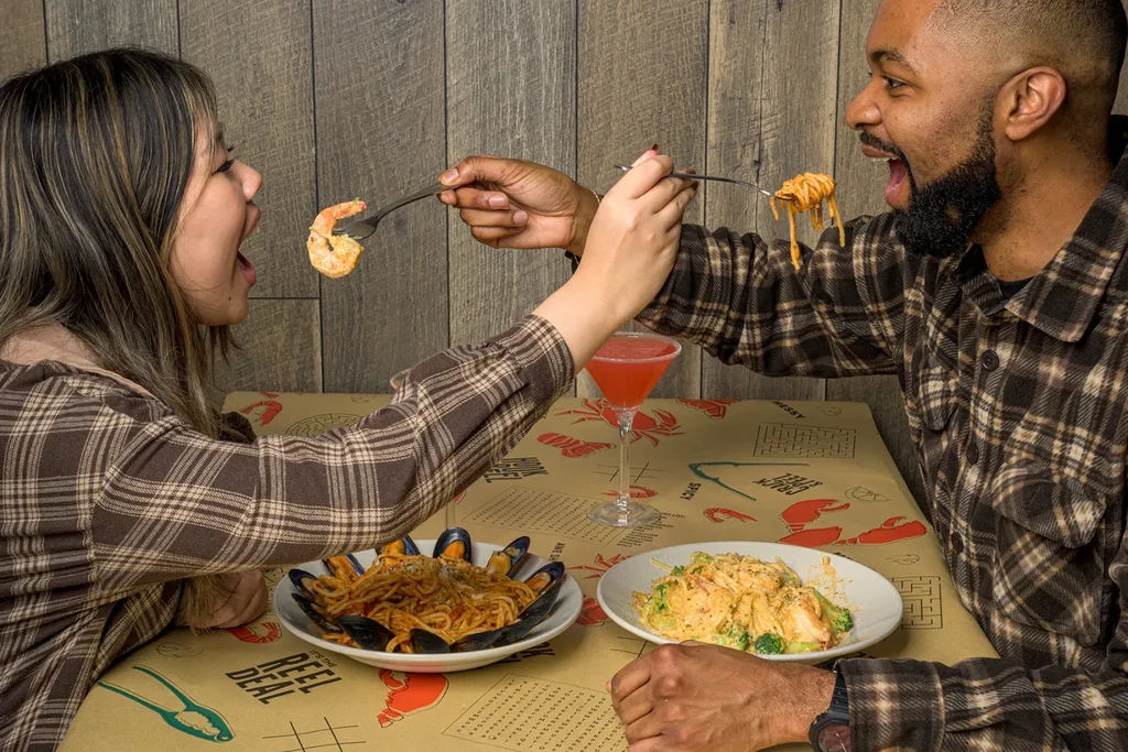 Man and woman playfully feeding each other shrimp and spaghetti.