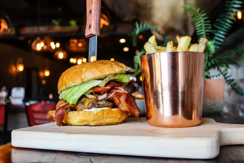 Gourmet burger with knife, french fries in cup on wooden board.