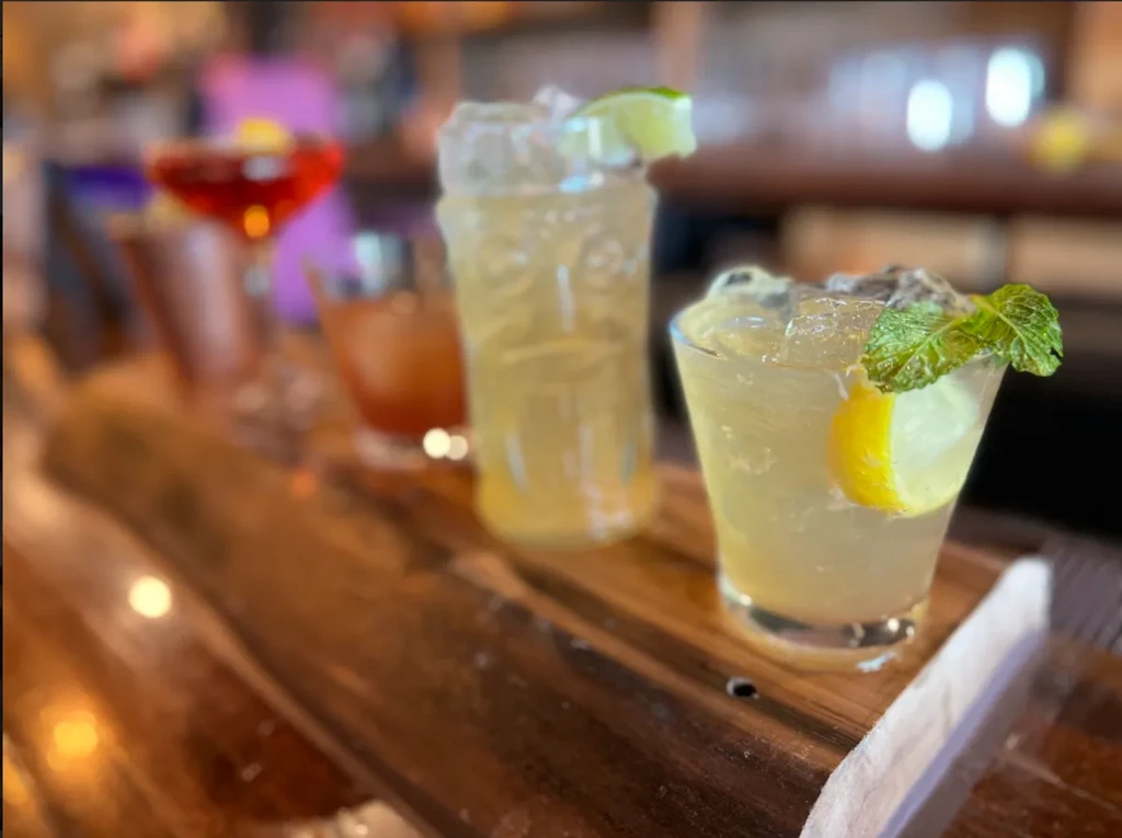 Multiple cocktails on a wooden board, two yellow drinks in focus.