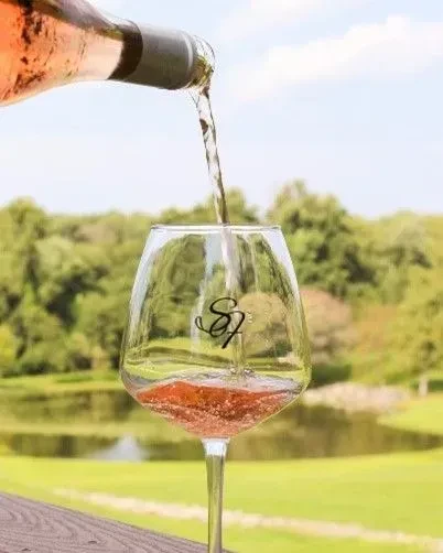 Rosé wine pours into an 'SJ' glass in a green outdoor setting.