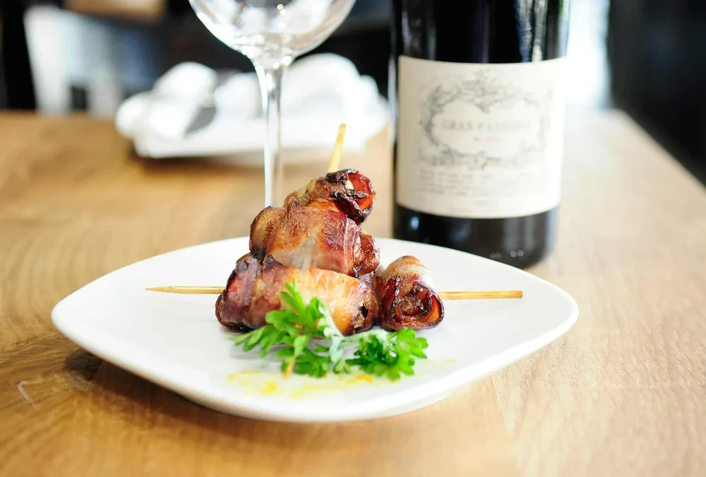 Four bacon-wrapped appetizers on skewers with parsley, sauce, and wine.