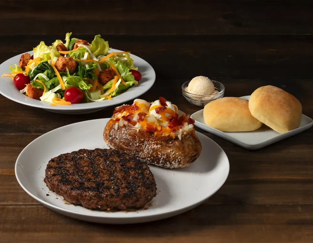 Grilled burger, loaded potato, salad, rolls, and butter on dark wood table.