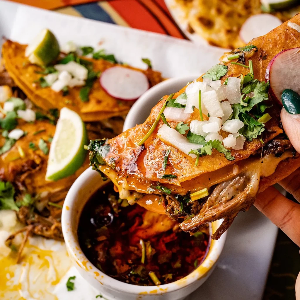 Golden-brown birria tacos, one being dipped into red consommé.