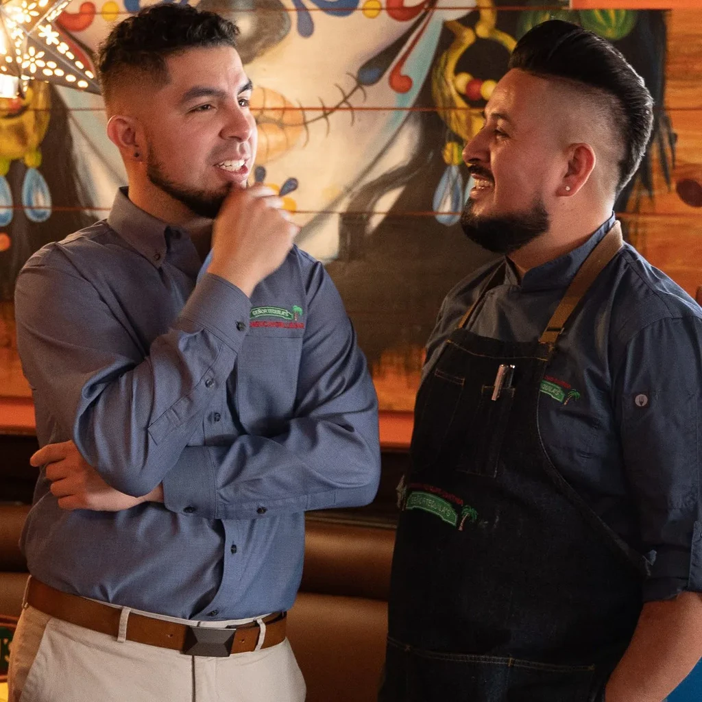 A chef and a man converse, smiling, against a colorful mural.