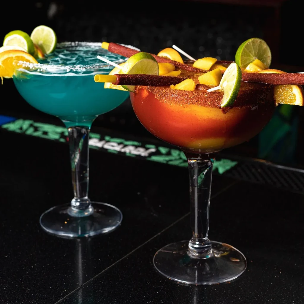 Elaborately garnished blue and mango-tajin cocktails on a dark bar top.