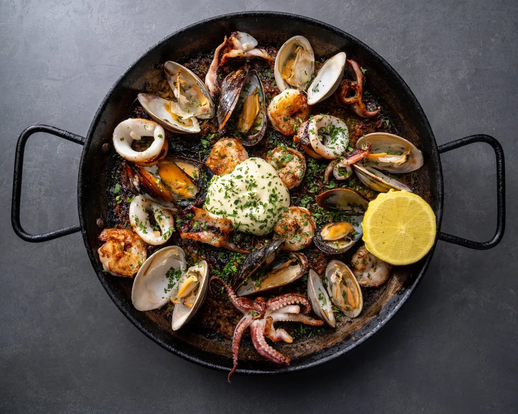 Cast-iron paella pan with seafood, dark rice, creamy sauce, and lemon.