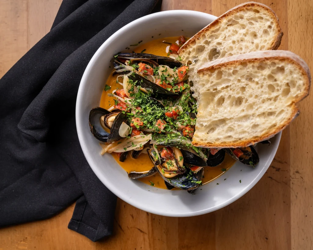 Mussels in orange broth with crusty bread on a wooden table.