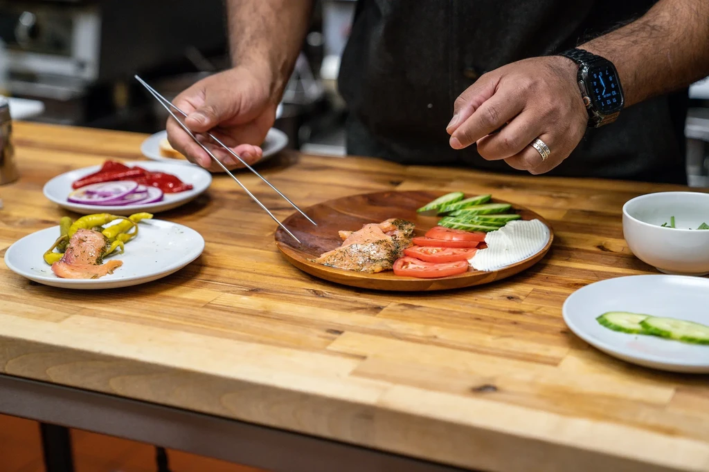 Dark-skinned hands meticulously plate smoked salmon, vegetables on wooden surface.