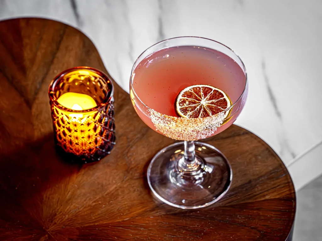 Reddish-pink cocktail with sugared rim, dried citrus, lit candle on wood.