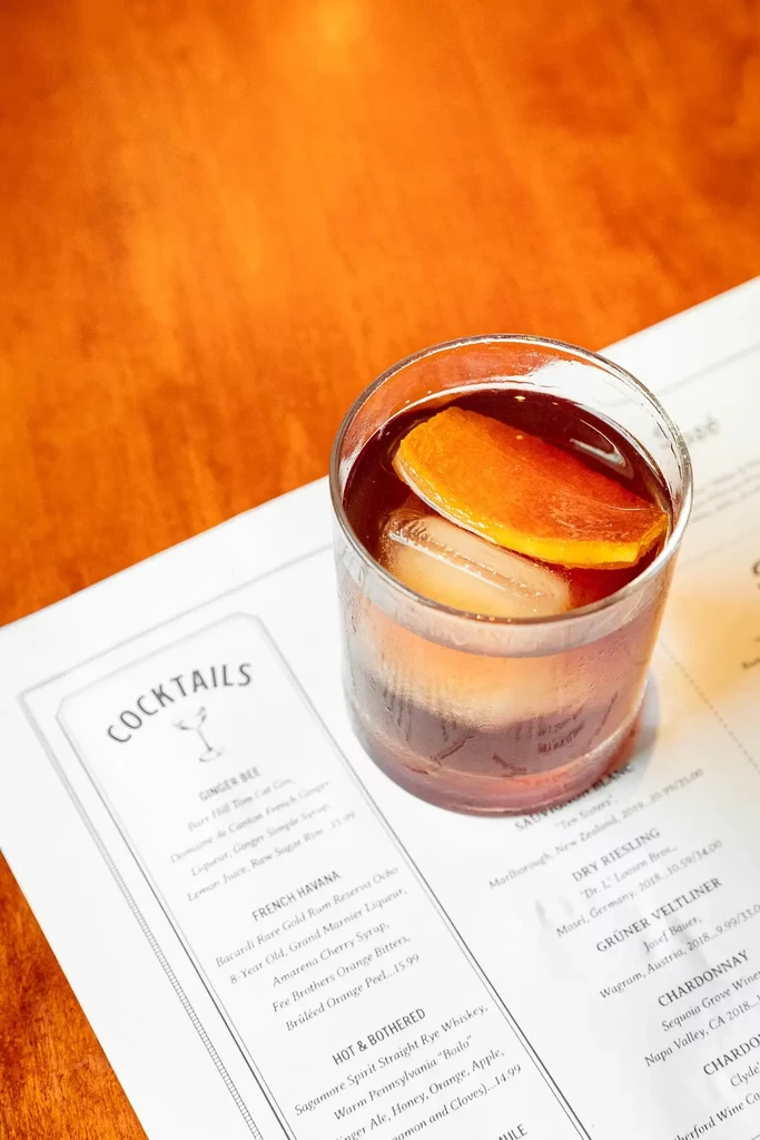 Amber cocktail with ice and orange garnish on a "COCKTAILS" menu.