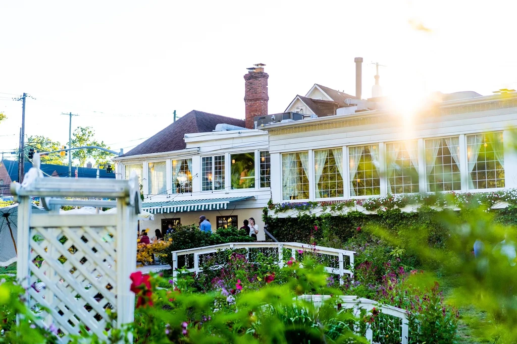 White restaurant building with patio, garden, and people in golden sunlight.