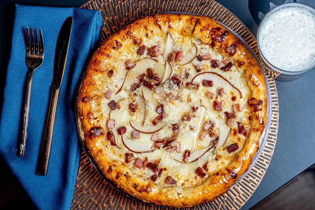 Golden-crusted pizza topped with cheese, meat, and pear, beside cutlery.