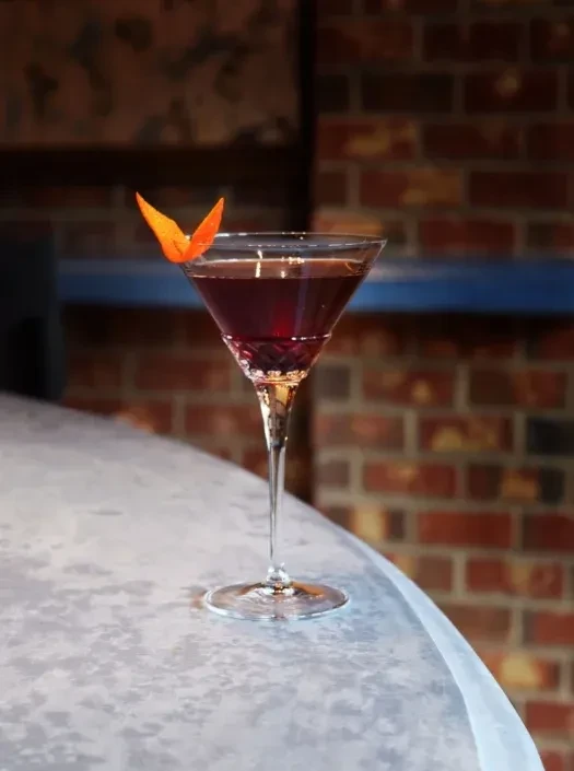 Reddish-purple cocktail with butterfly orange peel on bar, brick wall.