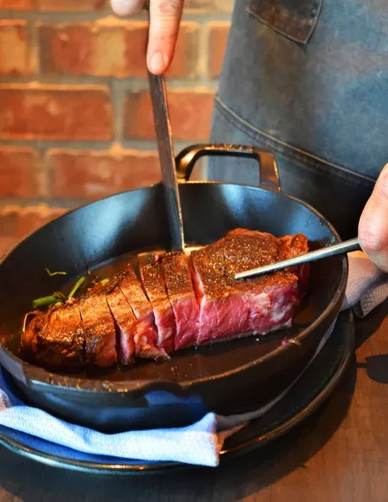 Close-up: Hands slice medium-rare steak in a cast-iron skillet with herbs.
