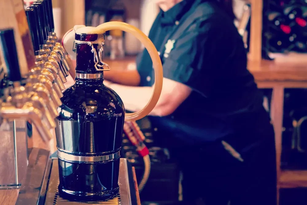 Dark growler bottle filling with foamy beer at a bar.