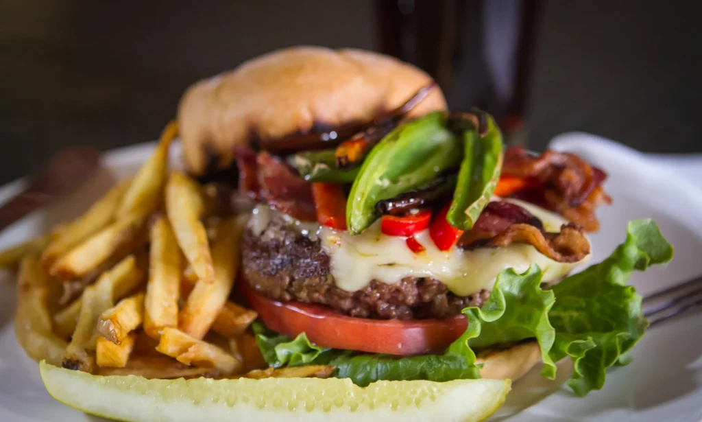 Gourmet burger with melted cheese, bacon, chili peppers, fries, pickle.