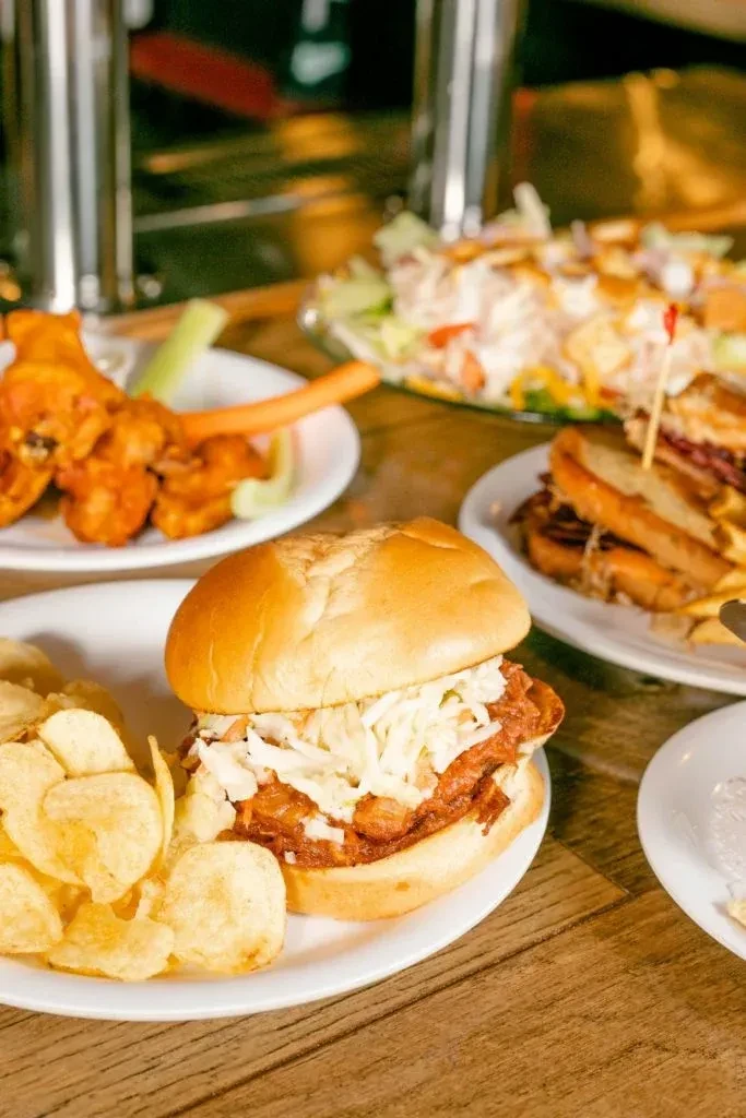 Pulled pork sandwich, buffalo wings, club sandwich, and salad on a wooden table.