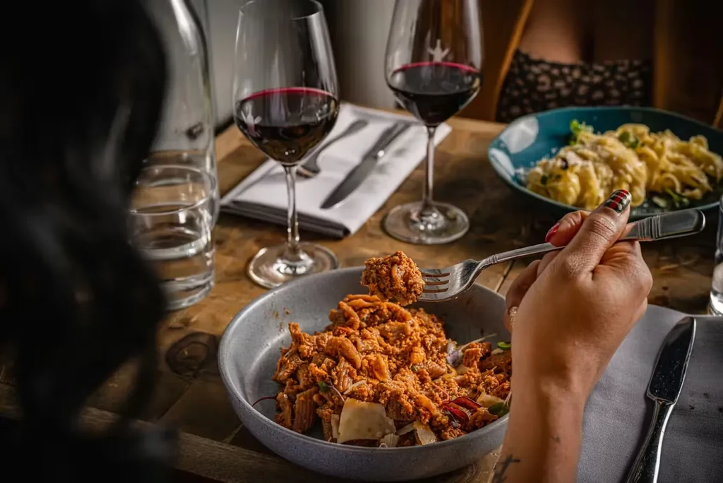Close-up of hand forking reddish pasta from gray bowl on table.