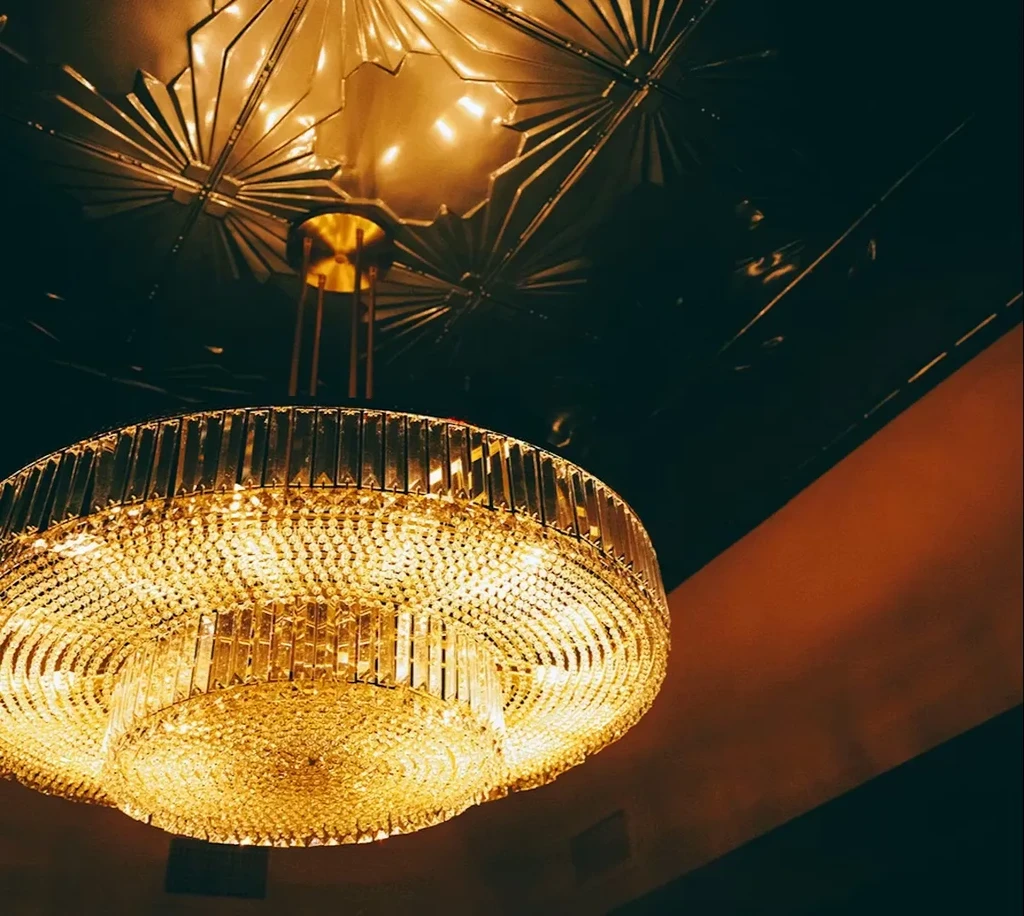 Multi-tiered crystal chandelier illuminates decorative Art Deco ceiling from below.