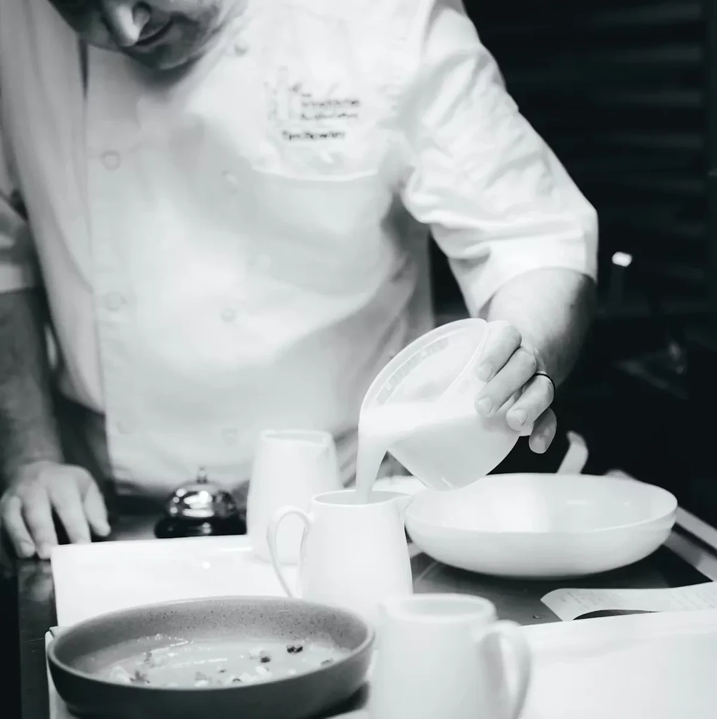 Black and white photo: Chef in jacket pours liquid for a dish.