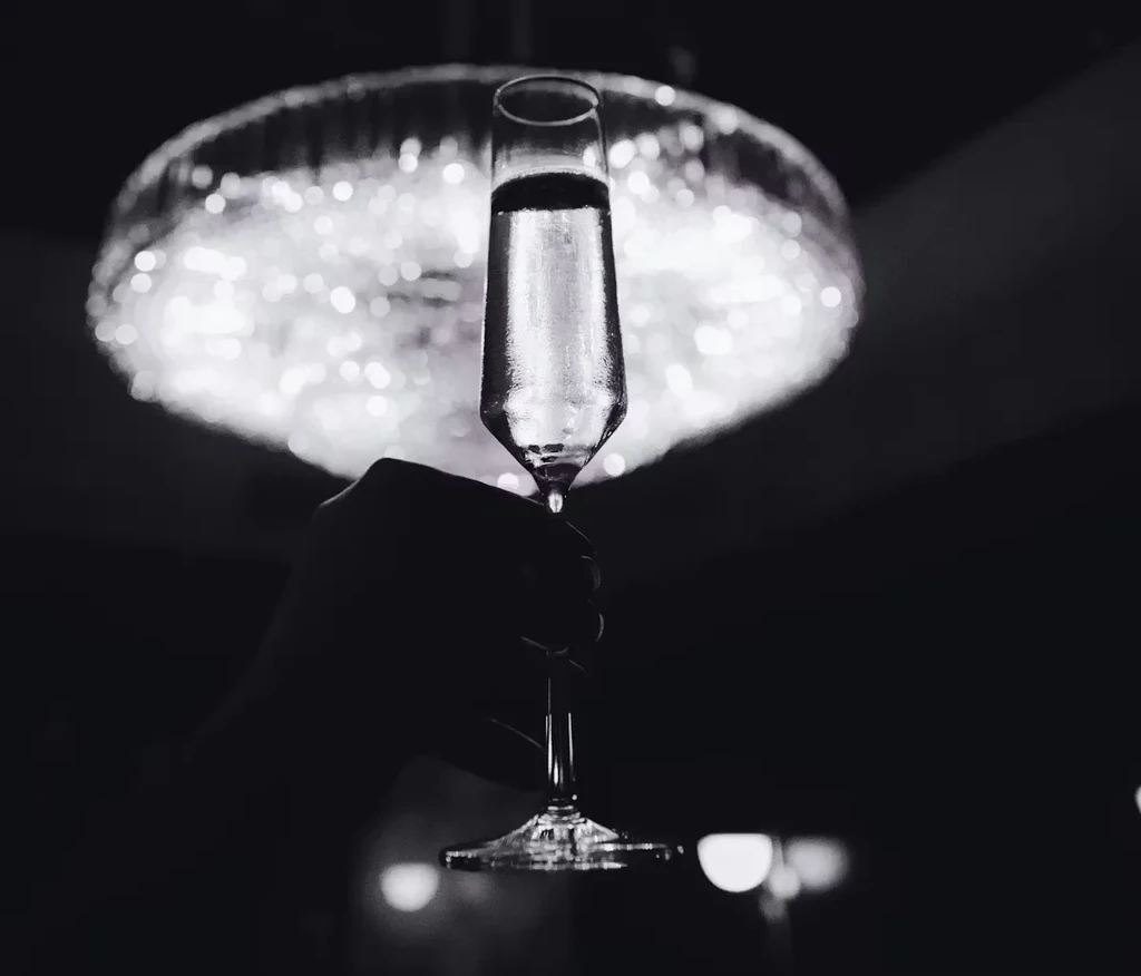 High-contrast B&W photo: Hand holds sparkling champagne flute, bokeh lights.