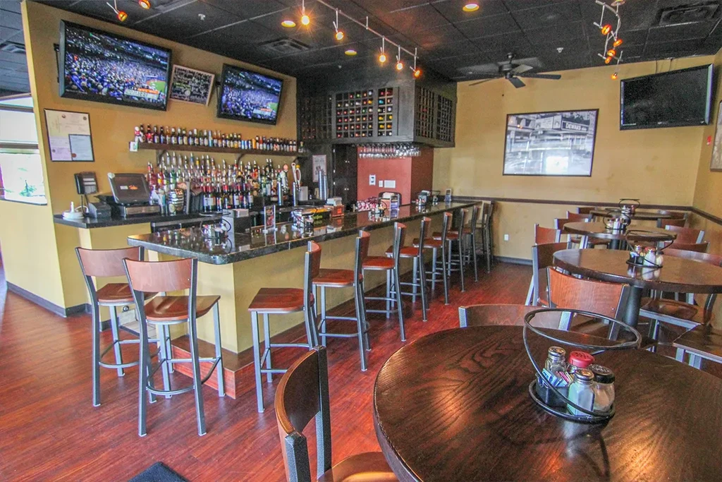 Empty sports bar interior featuring a stocked bar, dining tables, and TVs.