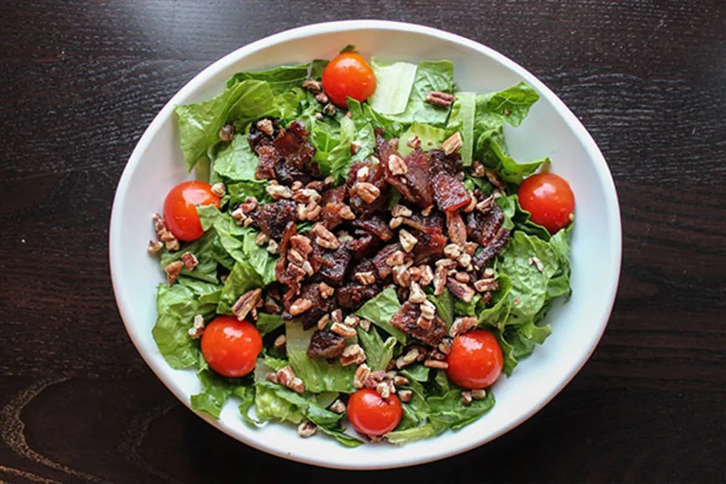 Green lettuce salad in white bowl with tomatoes, bacon, and pecans.
