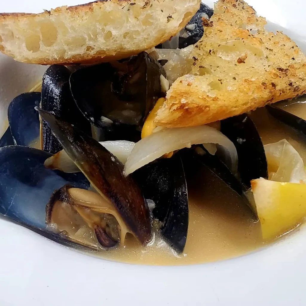Close-up of steamed mussels, sauce, lemon, onion, and herbed bread.