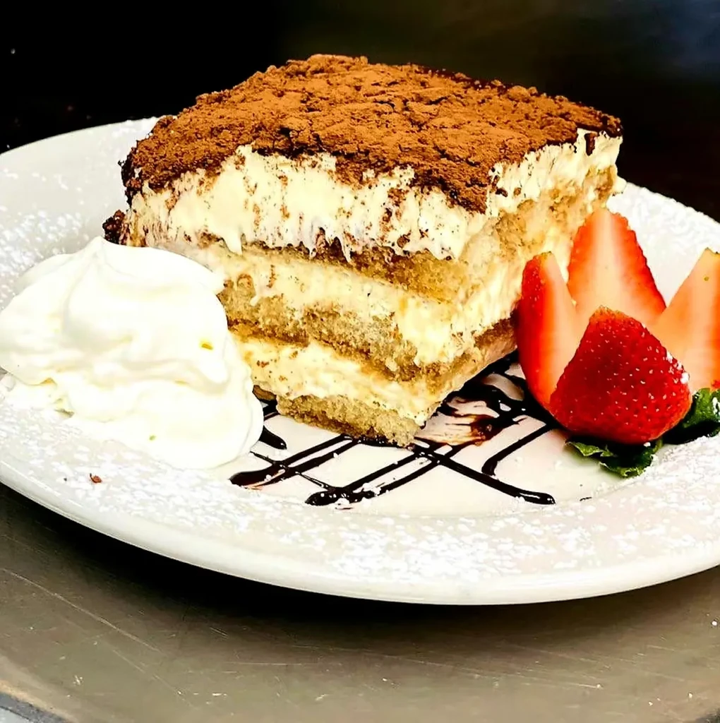 Tiramisu slice with cocoa, whipped cream, strawberries, chocolate syrup, powdered sugar.