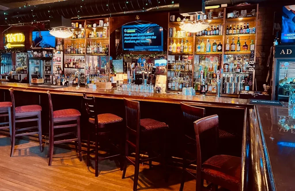 Interior of a well-stocked bar with two mounted televisions.