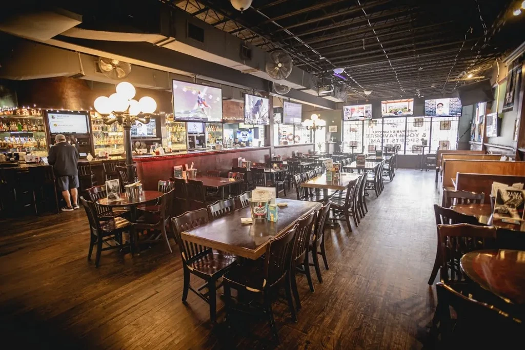 Dimly lit bar interior with wooden tables, chairs, and mounted TVs.
