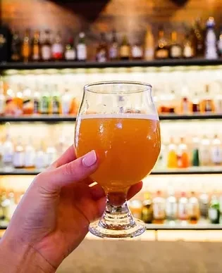 Hand holding amber beverage with foam against blurred bar background.
