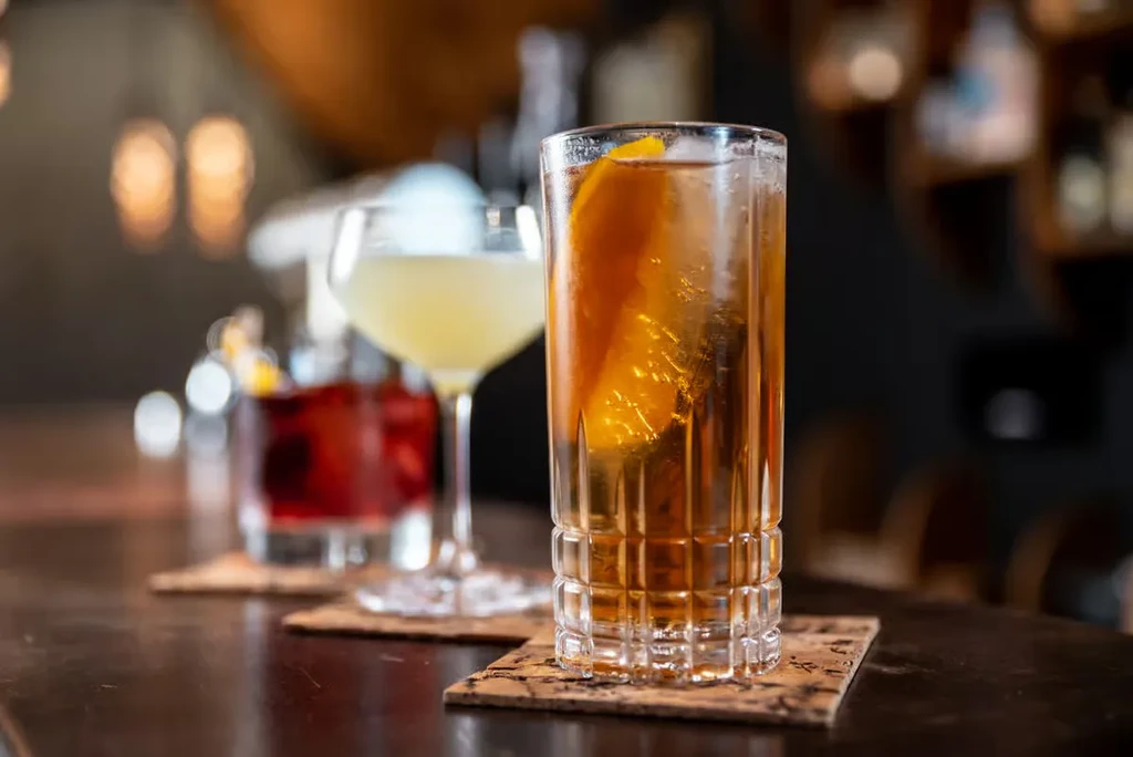 Three alcoholic cocktails on a dark bar with blurred lights.