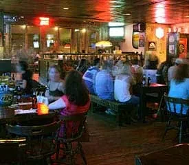Dimly lit bar interior with patrons, warm lighting, and wooden ceiling.