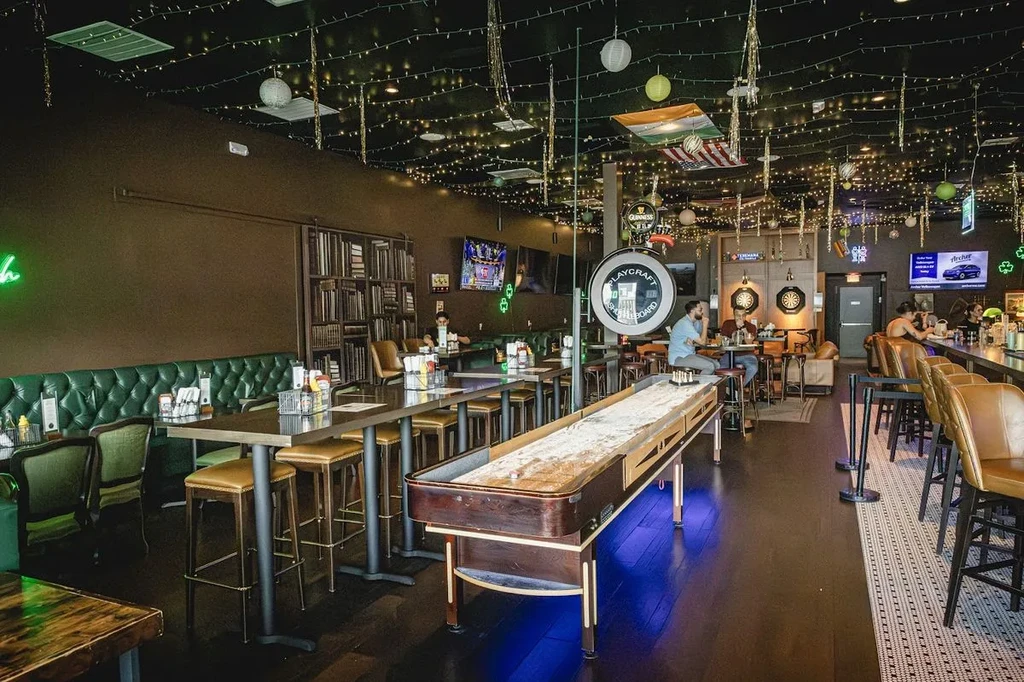Interior of a dimly lit bar with shuffleboard, dartboards, and string lights.