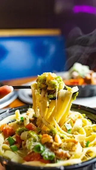 Fork lifts steaming pasta with pesto, tomatoes, and protein from bowl.
