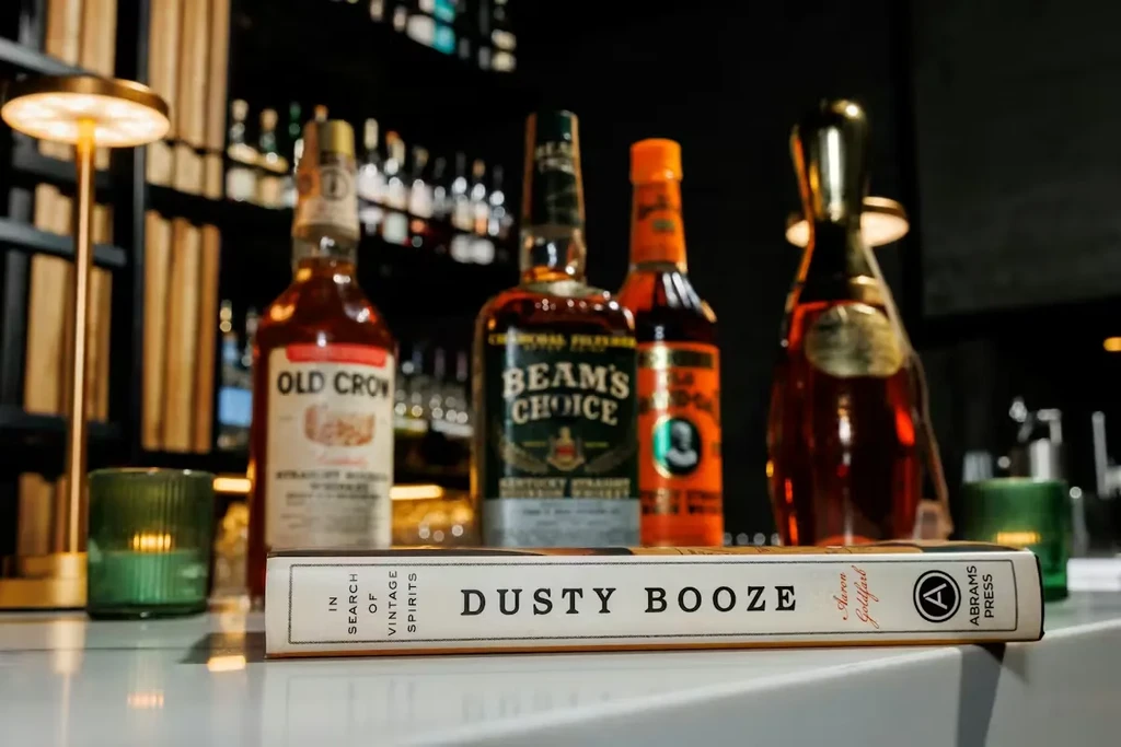 Five vintage whiskey bottles, a book, and a lamp on a bar.