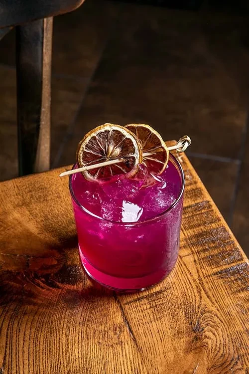 Fuchsia cocktail in a clear glass with ice and dried citrus.
