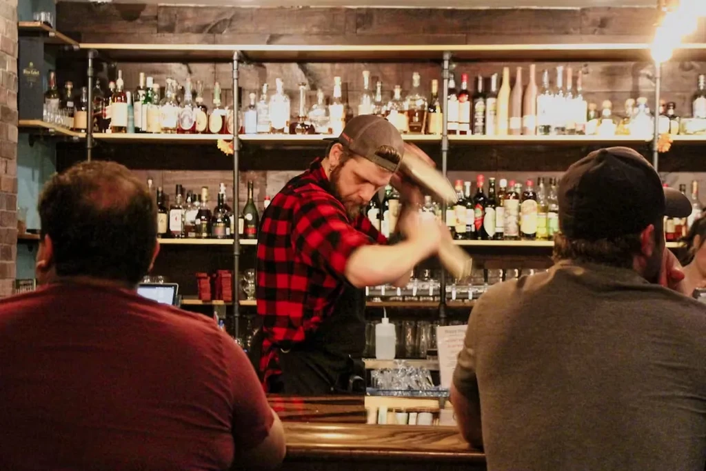 Bartender in plaid shirt shaking a cocktail shaker behind a bar.