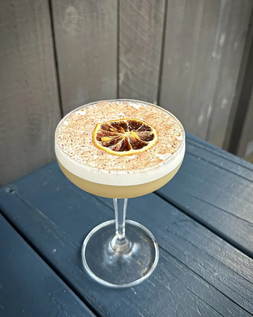 Creamy cocktail with foam, spice, and dried orange slice.