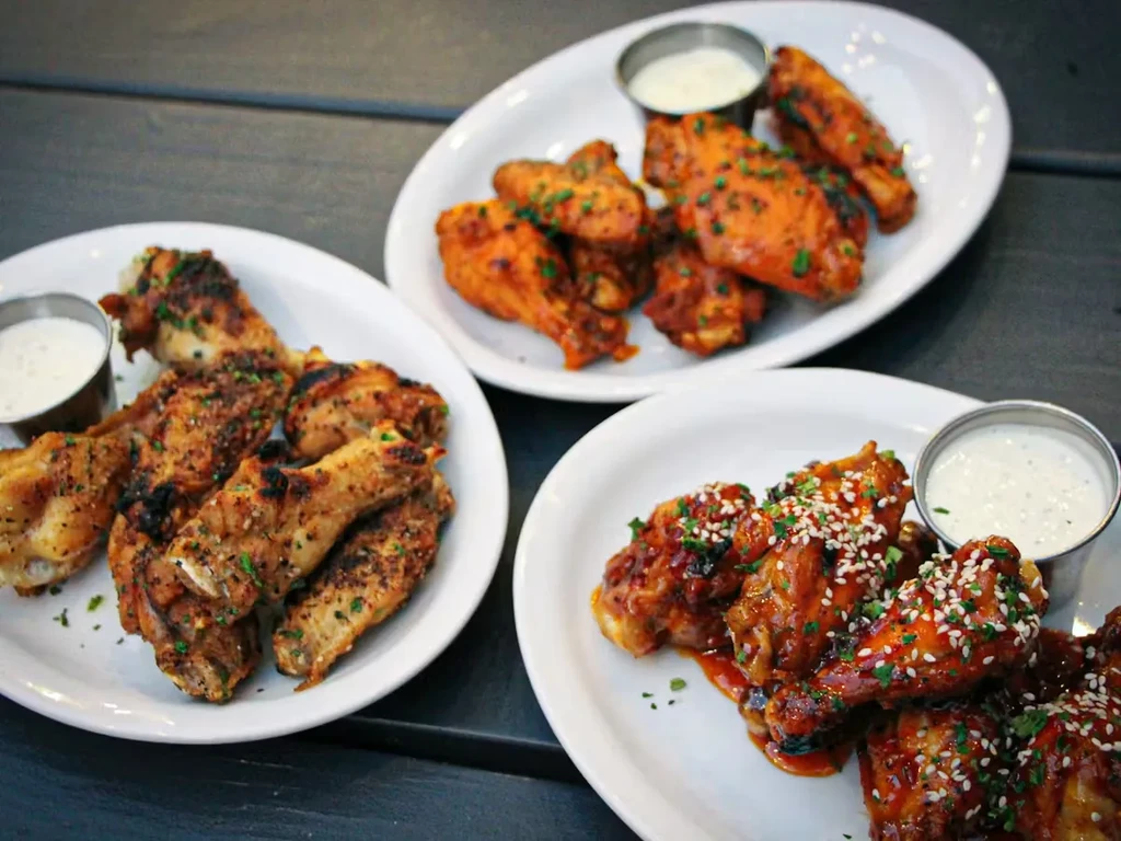 Three plates of chicken wings with dipping sauce on a dark table.