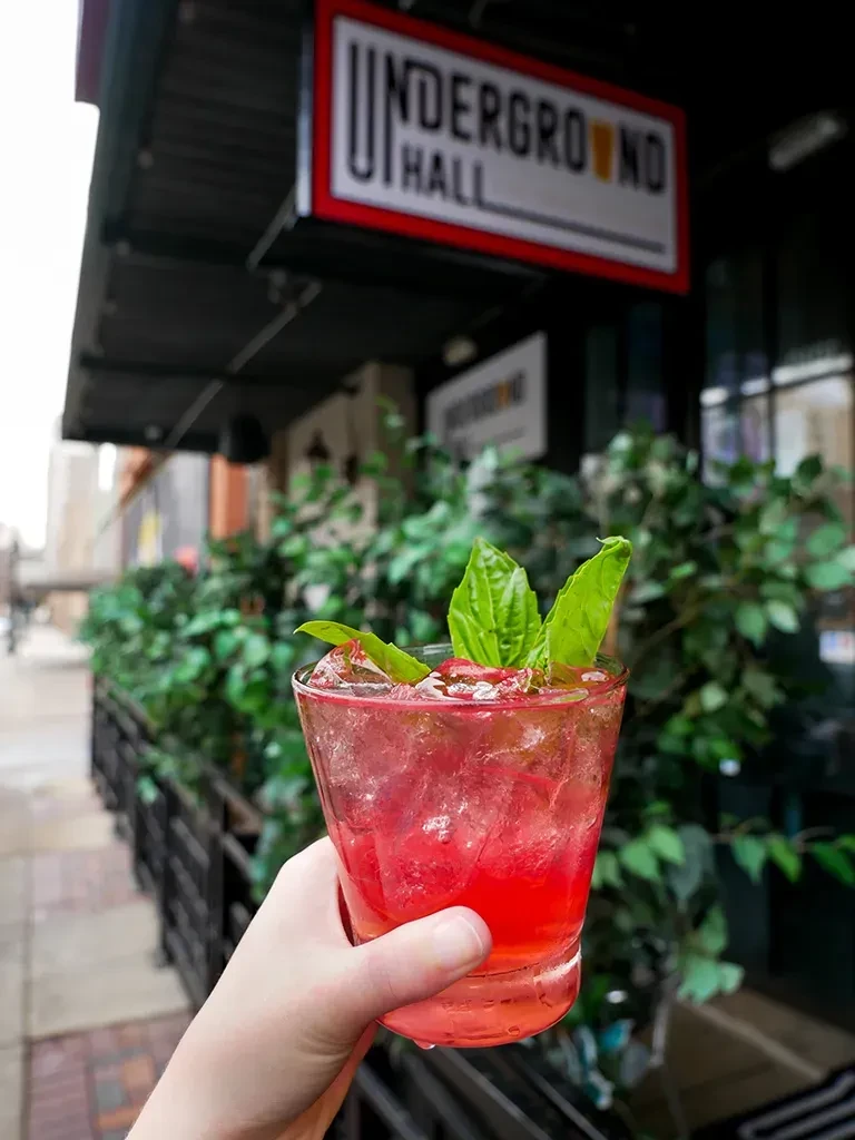 Close-up of hand holding pink cocktail with ice and basil outdoors.