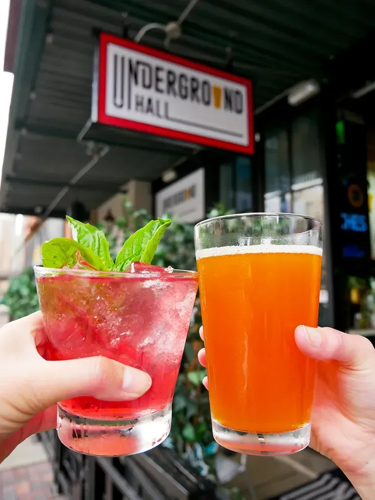 Two hands hold a red cocktail and an orange drink outdoors.