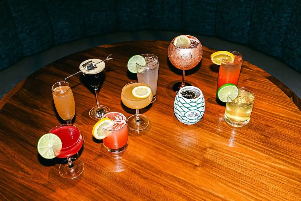 Nine distinct cocktails with various garnishes on a wooden table.