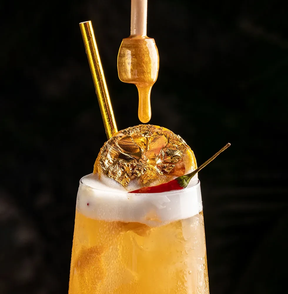 Close-up: Golden-yellow cocktail with gold leaf and chili garnish.