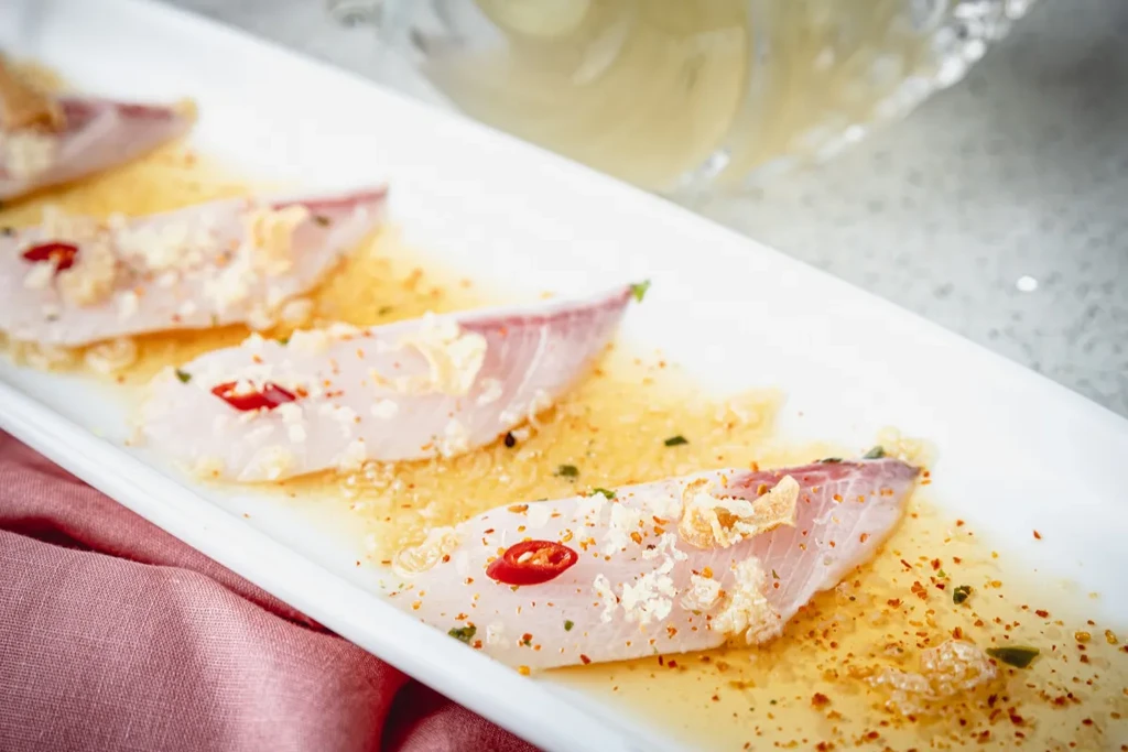 Four slices of fish with toppings in sauce on a white plate.