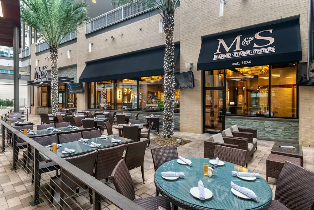 M&S restaurant patio with wicker tables, palm trees, and fairy lights.