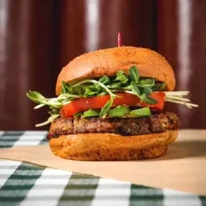 Gourmet burger with avocado, tomato, and sprouts on a checkered tablecloth.